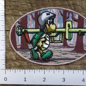 May include: Embroidered patch featuring a cartoon turtle wearing a helmet and carrying a large gun. The turtle is green and brown, and the gun is green and gray. The background is a brown and blue forest scene.