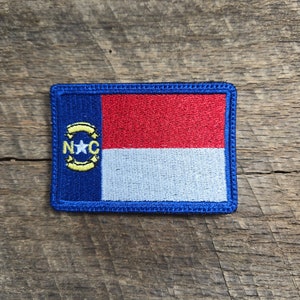 May include: A blue embroidered patch featuring the North Carolina state flag. The flag has a red and white stripe with a blue star and yellow letters "NC" in a circle.