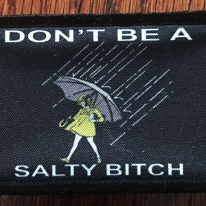 Könnte beinhalten: Schwarzer rechteckiger Aufnäher mit weißem Text "DON'T BE A SALTY BITCH". Der Aufnäher zeigt eine Illustration einer Person in einem gelben Regenmantel, die einen Regenschirm im Regen hält.