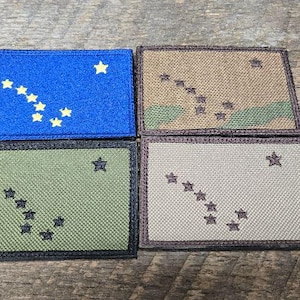 Op de afbeelding: Vier rechthoekige patches met het ontwerp van de vlag van de staat Alaska. De patches zijn in blauw, camouflage, olijfgroen en beige, elk met gele of beige sterren. De patches zijn waarschijnlijk voor kleding of uitrusting.