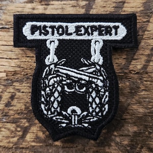 May include: A black and white embroidered patch featuring the text PISTOL EXPERT in a rectangular top bar. Below, a shield-shaped design displays two crossed pistols above a central circular emblem, all rendered in white thread on a black background.