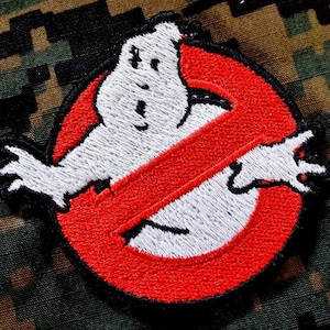 May include: A red and white embroidered patch with a ghost in the shape of a no symbol. The ghost is white and has a black outline. The patch is on a camouflage background.