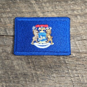 Pode incluir: Um remendo bordado azul com o selo do estado de Michigan. O selo apresenta um escudo com uma fita vermelha, dois alces e uma águia branca.