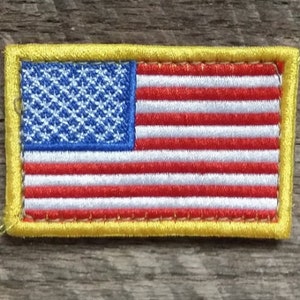 Può includere: Una piccola toppa ricamata della bandiera americana con un bordo giallo. La bandiera è rossa, bianca e blu.