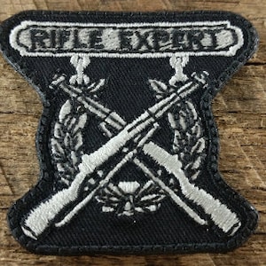 May include: A black embroidered patch with the text "Rifle Expert" in white. Two crossed rifles are embroidered in white with a wreath of leaves around them.