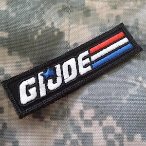 Puede incluir: Un parche bordado negro con las palabras "G.I. JOE" en letras blancas y una estrella azul. El parche tiene rayas rojas y azules.