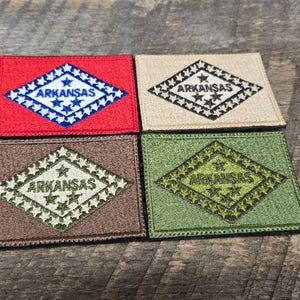 May include: Four square patches featuring the Arkansas state flag design. Each patch has a diamond shape with the word "ARKANSAS" and star details. The patches are in red, tan, brown, and green.