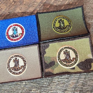 May include: Four rectangular patches with embroidered circular emblems. One patch is blue with a red and white emblem, the others are tan, green, and camouflage with a similar emblem. The patches are on a wooden surface.