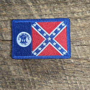 May include: A blue and red embroidered patch with the Confederate flag and the seal of the State of Georgia. The seal is a circle with the words "State of Georgia" and "1776" around the edge. The seal depicts a three-story building with a dome on top.