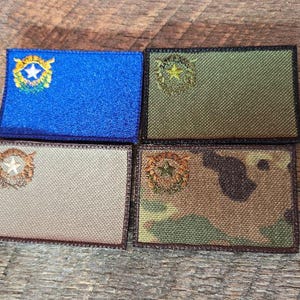 Könnte beinhalten: Vier rechteckige, gestickte Aufnäher in Blau, Olivgrün, Beige und Camouflage-Mustern. Jeder Aufnäher zeigt ein zentrales Emblem mit einem Stern und umgebenden Details. Die Aufnäher sind auf einer Holzoberfläche angeordnet.