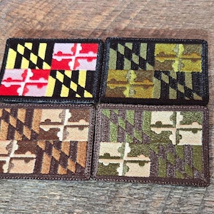 Puede incluir: Cuatro parches bordados con el diseño de la bandera del estado de Maryland. Los parches muestran los distintivos esquemas de color de la bandera: negro y dorado, rojo y blanco, y marrón y verde. Los parches son rectangulares con un borde cosido.