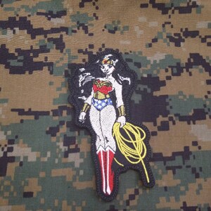 Può includere: Toppa ricamata raffigurante Wonder Woman nel suo costume classico, con una combinazione di colori rosso, oro, blu e bianco. La toppa ha un bordo nero e include un lazo giallo. Lo sfondo è un motivo mimetico.