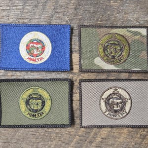 May include: Four rectangular patches with embroidered designs. One is blue with a circular emblem, the others are green, camouflage, and gray, each with a circular emblem. All have a black border and are on a wooden surface.