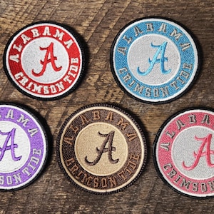 May include: Five circular embroidered patches with the text "ALABAMA CRIMSON TIDE" around the edge. Each patch features a large letter "A" in the center, with different color schemes: red, blue, purple, brown, and pink.