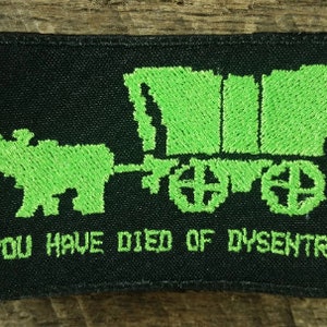Peut inclure: Un écusson brodé noir avec une image pixelisée verte d'un chariot couvert tiré par un bœuf. Le texte "YOU HAVE DIED OF DYSENTRY" est sous l'image.