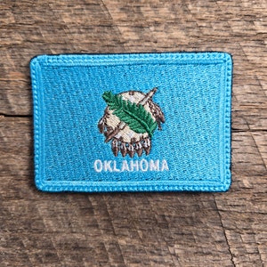 May include: A blue embroidered patch with the state seal of Oklahoma. The seal features a Native American headdress with feathers and a crossed tomahawk and peace pipe. The word "OKLAHOMA" is written in white below the seal.