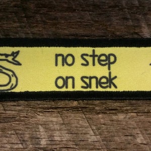 Pode incluir: Remendo amarelo com borda preta e o texto "no step on snek" com uma ilustração de cobra preta de cada lado.
