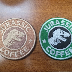 Puede incluir: Dos parches redondos bordados con una silueta de dinosaurio y el texto "Jurassic Coffee". Un parche es marrón con texto blanco y el otro es verde con texto blanco.