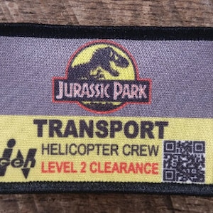 Pode incluir: Um remendo preto e amarelo com o texto "Jurassic Park" e um logotipo de dinossauro. O remendo também diz "Transport Helicopter Crew Level 2 Clearance" e tem um código QR.