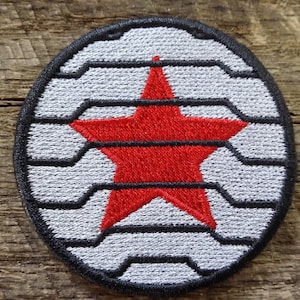 Peut inclure: Un écusson brodé blanc et noir avec une étoile rouge au centre. L'étoile est à cinq branches et a un contour blanc. L'écusson est circulaire et a une bordure noire.