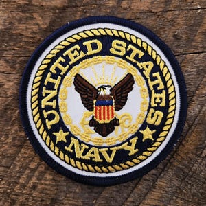 Puede incluir: Un parche bordado circular con el emblema de la Marina de los Estados Unidos. El diseño incluye un águila central con las alas extendidas, un escudo y un borde de cuerda dorada sobre un fondo blanco con el texto azul oscuro UNITED STATES NAVY.