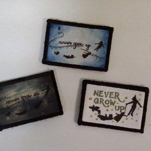May include: Three embroidered patches with a black border and a white background. The patches feature the text "never grow up" and a silhouette of Peter Pan flying with the Lost Boys. The patches are different sizes and have different designs.