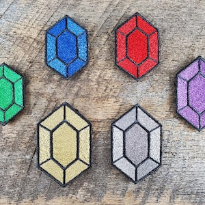 Puede incluir: Seis parches bordados con forma de gema y contorno negro. Las gemas son de color azul, rojo, verde, morado, dorado y gris.