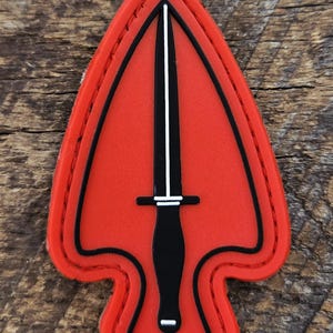 May include: A shield-shaped patch in bright red with a black border and a central black dagger graphic. The dagger features a thin white vertical line running down the blade and white horizontal accents on the crossguard and the base of the handle.