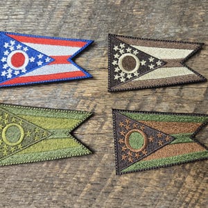 Può includere: Quattro toppe ricamate a forma di bandiera dell'Ohio. Una toppa è blu, rossa e bianca. Le altre tre toppe sono in tonalità di marrone e verde. Le toppe sono su una superficie di legno.