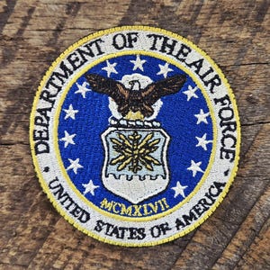 Op de afbeelding: Een ronde geborduurde patch met het embleem van het Department of the Air Force. Het toont een bruine adelaar boven een schild met een geel stervormig embleem, omringd door witte sterren op een blauw veld, met de tekst MCMXLVII onderaan.