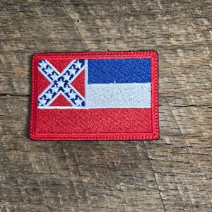 Pode incluir: Um remendo bordado com a bandeira confederada do estado do Mississippi. O remendo é vermelho, branco e azul e está sobre um fundo de madeira.