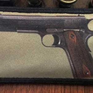 May include: A rectangular patch featuring a detailed image of a handgun. The gun is depicted in shades of brown and black, with a wooden grip. The patch has a black border and a tan background.