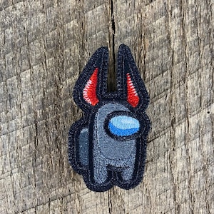 Puede incluir: Un parche bordado gris de un personaje de Among Us con cuernos rojos y una visera azul.