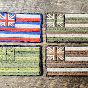 Puede incluir: Cuatro parches rectangulares bordados con el diseño de la bandera hawaiana. Un parche tiene las tradicionales rayas rojas, blancas y azules con una Union Jack en la esquina. Los otros tres parches tienen un diseño similar pero con diferentes combinaciones de colores, incluyendo tonos de verde y marrón.