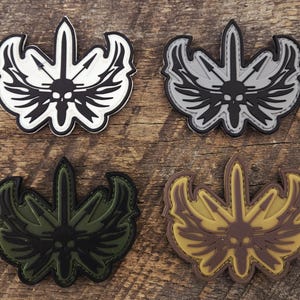 May include: Four tactical morale patches featuring a central skull design with wings and a vertical sword. The patches are rendered in four distinct color combinations: white and black, gray and black, olive green and black, and tan and brown.