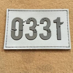以下が含まれることがあります： 白い長方形のパッチで、ステッチの縁取りがあり、灰色の数字「0331」が印刷されています。