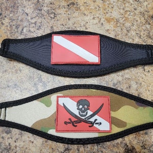 Può includere: Due cinturini per maschera da sub con design diversi. Un cinturino è nero con una bandiera da sub rossa e bianca. L'altro cinturino è mimetico con una bandiera da sub rossa e bianca con un teschio e ossa incrociate nere.
