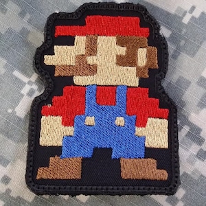Puede incluir: Un parche bordado pixelado del icónico personaje de videojuego, Mario. El parche presenta a Mario con su característico mono rojo y sombrero, con bigote y barba marrones. El parche está hecho de tela negra con bordado de colores.