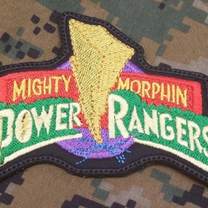 Könnte beinhalten: Ein gestickter Aufnäher mit den Worten "Mighty Morphin Power Rangers" in Grün, Gelb, Rot und Weiß und einem Blitz-Design.