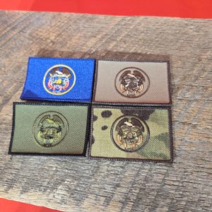 Puede incluir: Cuatro parches rectangulares bordados con emblemas. Uno es azul con un emblema dorado, otro es beige con un emblema dorado, un tercero es verde oliva con un emblema dorado, y el último es camuflaje con un emblema dorado. Los parches están sobre una superficie de madera.