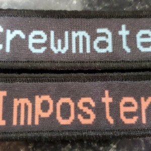 May include: Two black fabric patches with white stitching around the edges. The top patch has the word "Crewmate" in light blue pixelated text. The bottom patch has the word "Imposter" in orange pixelated text.