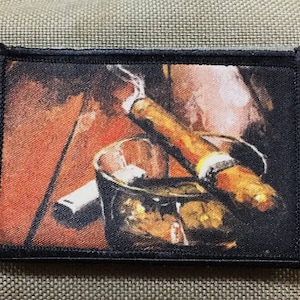 May include: A close-up of a brown and black cigar resting in a glass of amber liquid. The cigar is partially lit and smoke is rising from it. The glass is on a wooden surface.