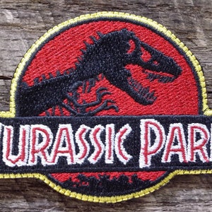 Puede incluir: Un parche bordado negro y rojo con una silueta de dinosaurio y el texto "Jurassic Park" en letras blancas.