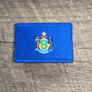May include: A blue embroidered patch featuring the state seal of Connecticut. The seal depicts a blue shield with a white pine tree, a white eagle, and two white figures.