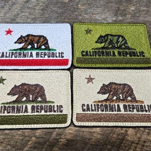 Puede incluir: Cuatro parches rectangulares con el diseño de la bandera de la República de California. Cada parche muestra un oso, una estrella y el texto "CALIFORNIA REPUBLIC". Los parches tienen diferentes combinaciones de colores: blanco con rojo, verde y marrón.