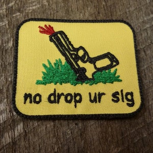 Op de afbeelding: Een gele geborduurde patch met een zwart-witte cartoon-pistool met een rode vlam die uit de loop komt. Het pistool ligt op groen gras. De tekst "no drop ur slg" staat in het zwart onder het pistool.