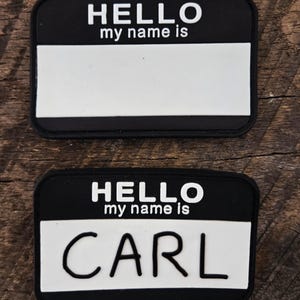 May include: Two rectangular name tag patches with a black border and background. The top portion features the white text HELLO my name is, while the bottom portion is a white rectangular space. The bottom patch has the name CARL written in black.