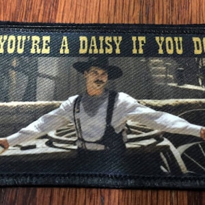May include: A black and white embroidered patch featuring the text "You're a daisy if you do" and a scene from a Western film. The scene depicts a man in a white shirt and black hat holding a gun.