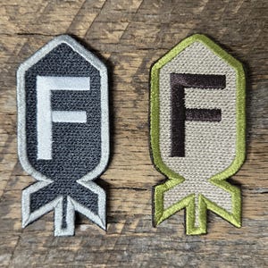 May include: Two embroidered patches. One is black with a white "F" and silver trim. The other is tan with a brown "F" and green trim. Both patches feature a shield-like design with the letter "F" and a stylized base.