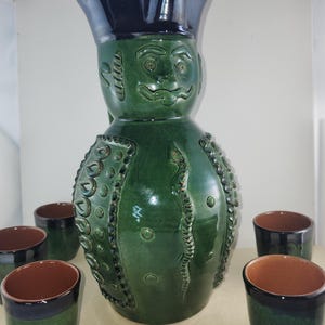 May include: A green ceramic jug with a face and a snake design, and six matching green and brown ceramic cups.